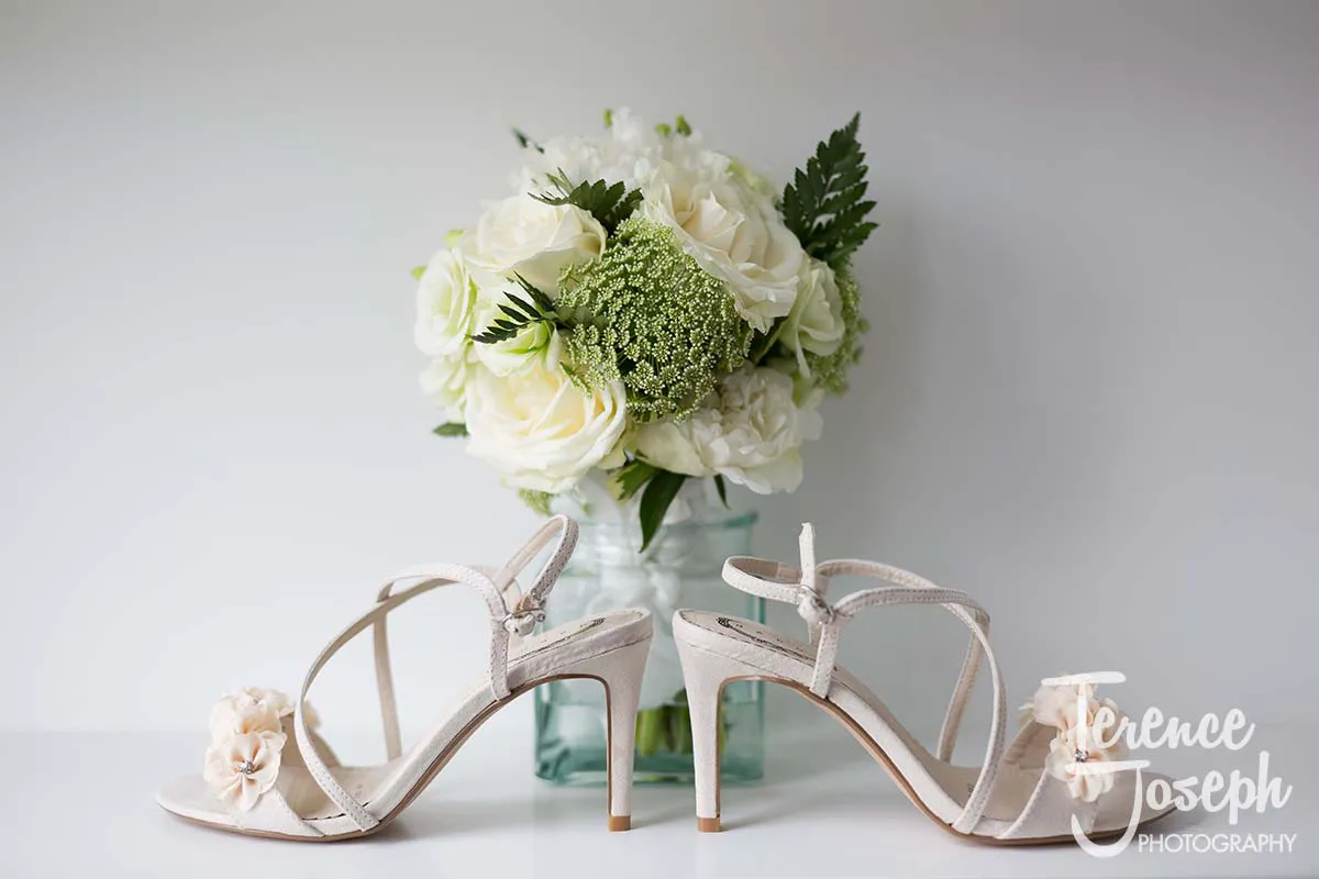 01_County_Arms_Wandsworth_Wedding_Bridal_Prep_Photos White high-heeled sandals with floral accents are placed in front of a bouquet of white roses and greenery in a glass vase, capturing the elegance of a County Arms Wandsworth wedding. The background is a soft, neutral tone.