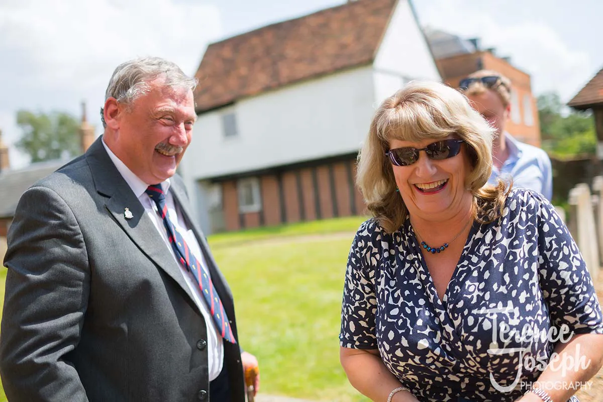 01_Moreteyne_Manor_Wedding_Church_Wedding A man in a suit with a striped tie and a woman in a patterned blouse stand outdoors, smiling and laughing. Captured by Moreteyne Manor Wedding Photographer, the scene is set against a picturesque park with lush grass, trees, and charming buildings in the background.