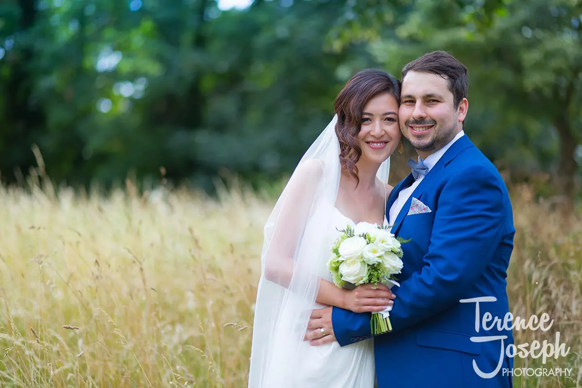 02_Terence_Joseph_Photogarapher_MJ A newlywed couple stands in a field of tall grass near County Arms Wandsworth, smiling warmly. The bride wears a white dress and holds a bouquet of white flowers, while the groom is in a blue suit. They appear happy and relaxed, enjoying their wedding moment together outdoors.