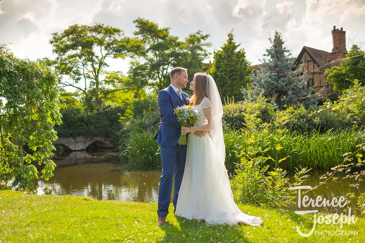 02_Terence_Joseph_Photography_BP A bride and groom stand embracing in a lush garden by a pond at Moreteyne Manor. The groom wears a blue suit while the bride, in her white dress with a veil, holds a bouquet. Trees and a traditional brick house enhance this picturesque scene under the partly cloudy sky, beautifully captured by the wedding photographer.