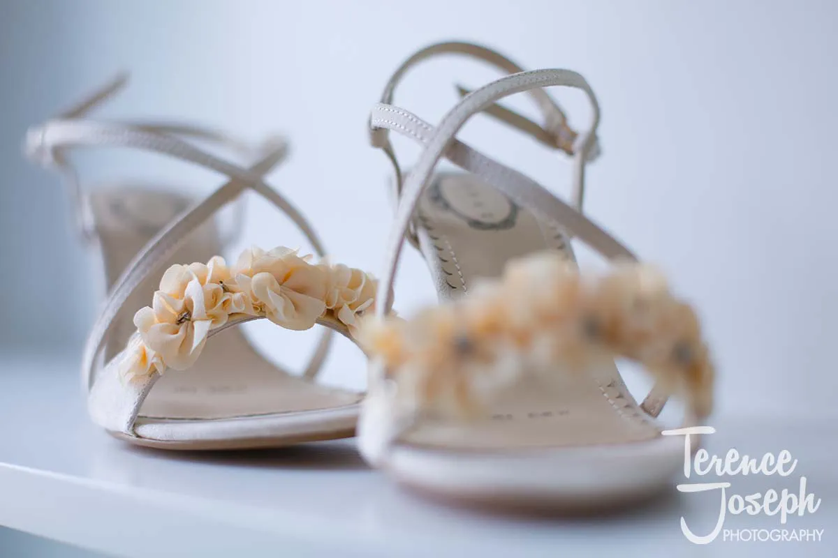 03_County_Arms_Wandsworth_Wedding_Bridal_Prep_Photos A pair of elegant white high-heeled sandals with floral accents on the straps, perfect for a County Arms Wandsworth wedding, placed on a pristine white surface.