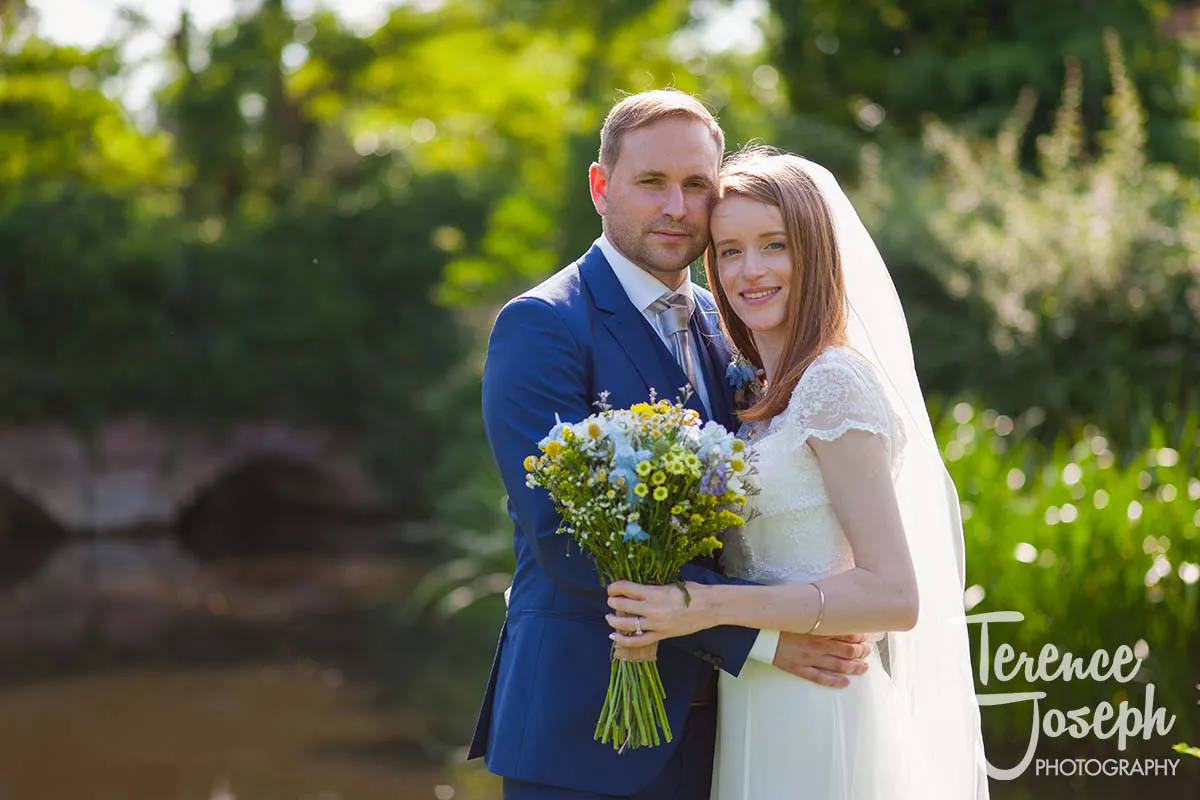 03_Terence_Joseph_Photography_BP In a lush garden at Moreteyne Manor, a bride and groom pose together with a small pond visible in the background. The bride holds a bouquet of wildflowers, and both are smiling gently at the camera, captured beautifully by their wedding photographer.