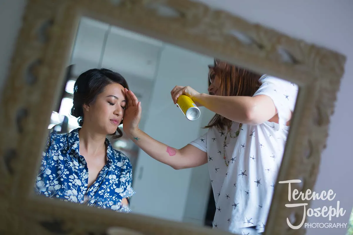 04_County_Arms_Wandsworth_Wedding_Bridal_Prep_Photos A woman getting her makeup done for a County Arms Wandsworth wedding is reflected in a mirror. She wears a blue floral shirt and has her eyes closed. Another person, partially visible, applies makeup with a brush and holds a spray can. A decorative frame surrounds the mirror.