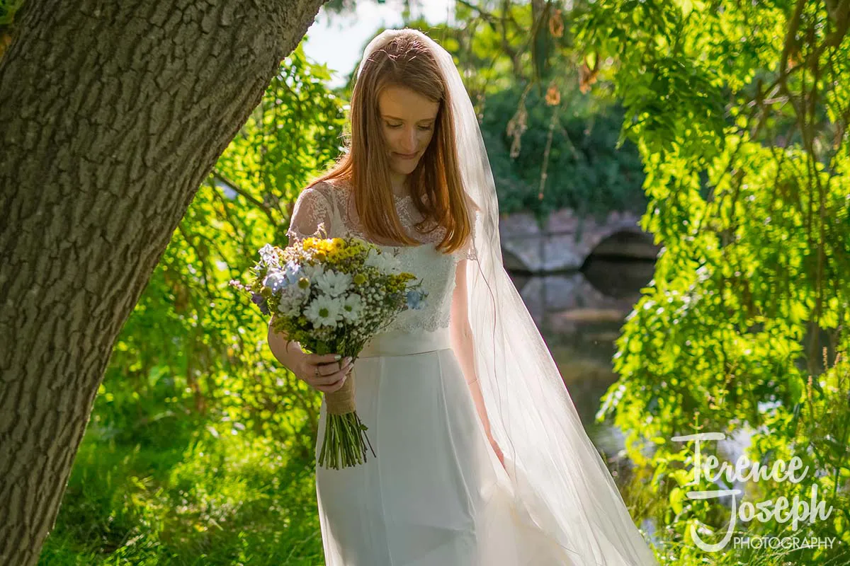 04_Terence_Joseph_Photography_BP A bride in a white dress and veil stands under a tree, holding a bouquet of flowers. Sunlight streams through the leaves, creating a serene and romantic atmosphere. Captured by Moreteyne Manor Wedding Photographer, a bridge and greenery enhance the enchanting backdrop.