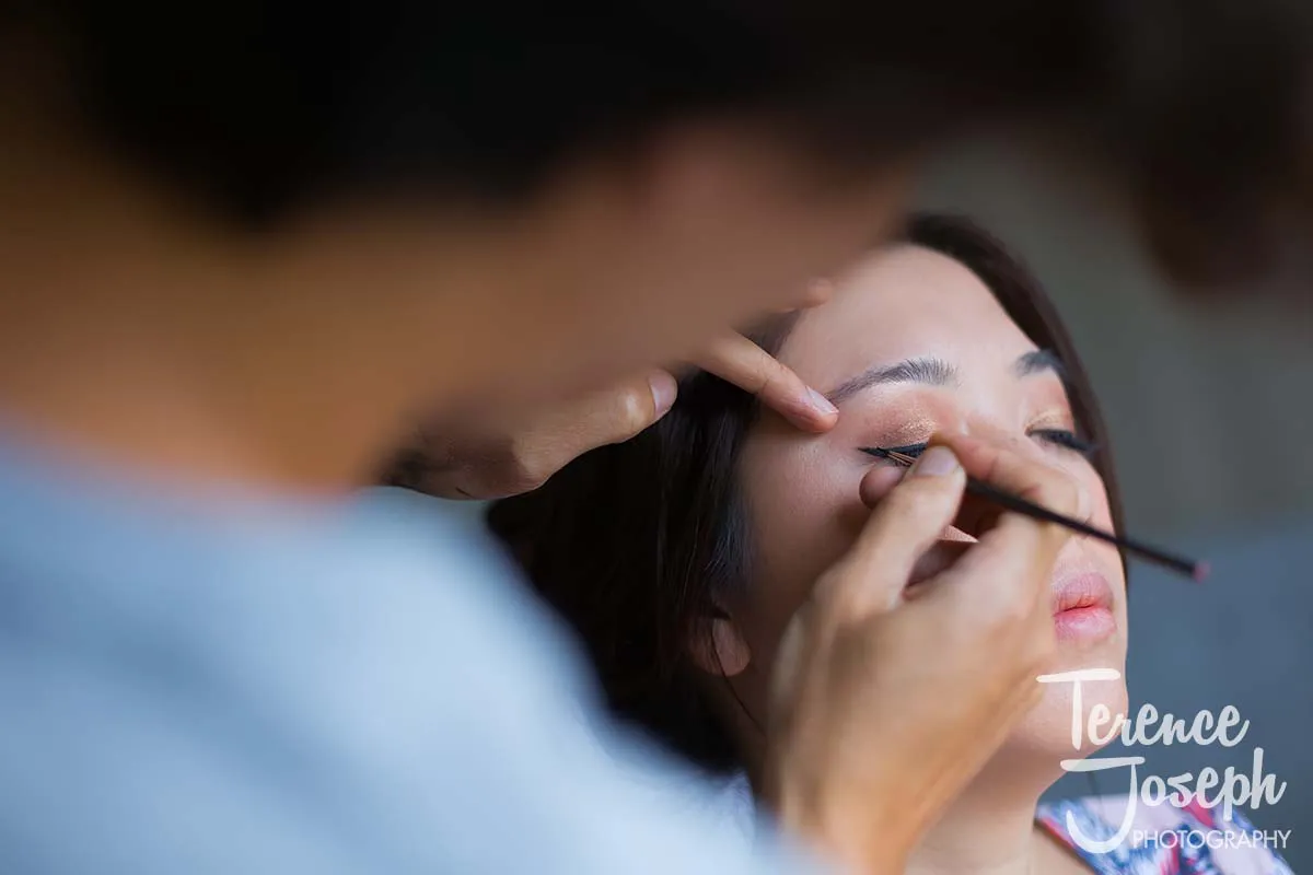 05_County_Arms_Wandsworth_Wedding_Bridal_Prep_Photos A person is having eyeliner applied to their eyelid with a brush, as another holds it steady. The scene captures the intricate art of makeup, reminiscent of the elegance seen at a County Arms Wandsworth wedding. The image includes a watermark for "Terence Joseph Photography.