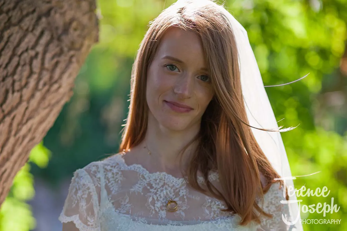 05_Terence_Joseph_Photography_BP A bride with long reddish-brown hair stands outdoors, smiling gently at her Moreteyne Manor wedding. She is wearing a white lace dress and veil, with green leaves and sunlight painting a serene backdrop.