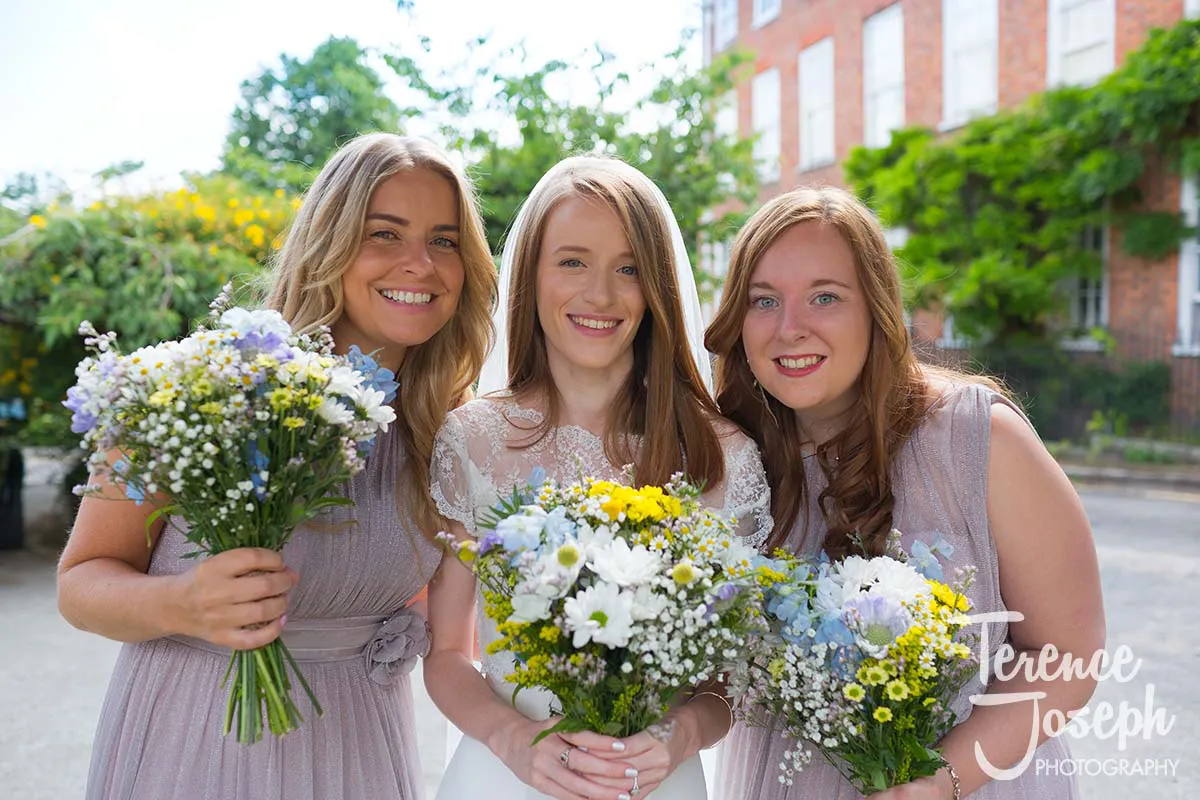 07_St_Andrews_Church_Wedding_Ceremony Three women smile outdoors in light purple dresses, perfectly captured by a Moreteyne Manor Wedding Photographer. Two hold bouquets of wildflowers featuring daisies and yellow accents, set against lush greenery and a charming brick building backdrop.