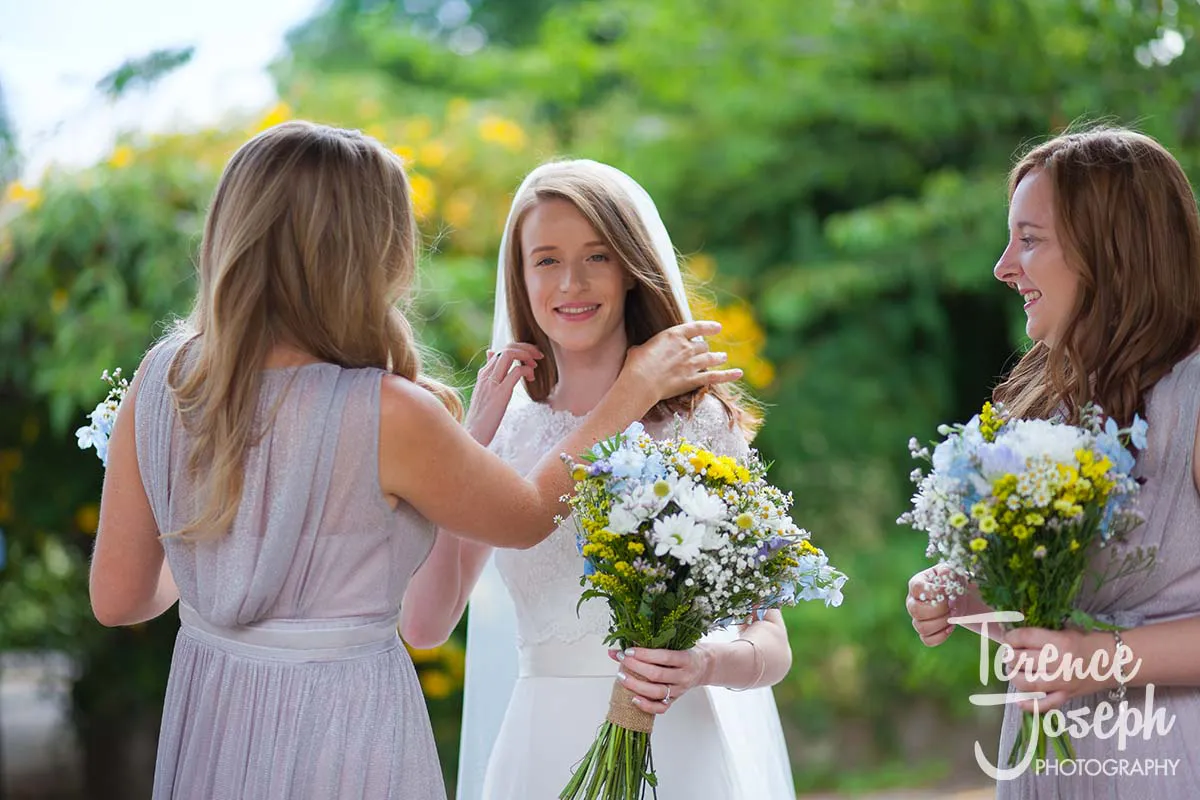 08_St_Andrews_Church_Wedding_Ceremony In a picturesque garden setting, captured by a Moreteyne Manor wedding photographer, three women gracefully pose. Two in light gray dresses hold bouquets of yellow and white flowers, while one adjusts the smiling bride's hair. A backdrop of lush greenery completes this enchanting scene.