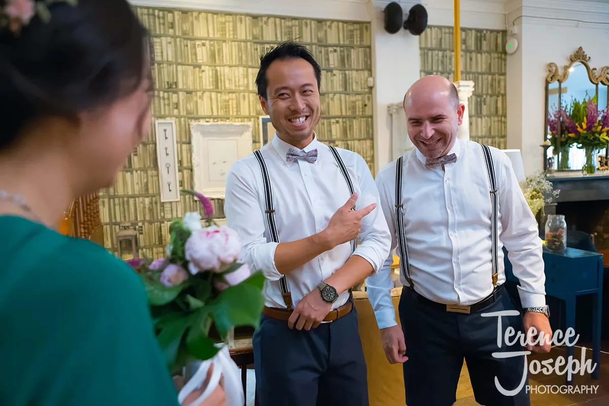 09_County_Arms_Wandsworth_Wedding_Ceremony_Photos Three people are at a gathering, possibly a County Arms Wandsworth wedding. Two men in white shirts, bow ties, and suspenders smile as one gestures. A person with a bouquet stands in the foreground facing them. In the background, a bookshelf and mirror complete the scene.
