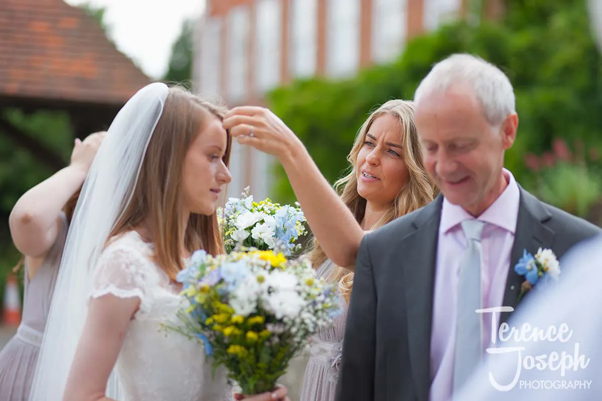 09_St_Andrews_Church_Wedding_Ceremony A bride holds a bouquet of white and yellow flowers as a woman adjusts her veil, captured beautifully by a Moreteyne Manor Wedding Photographer. Nearby, a man in a suit stands smiling under the sunny sky.