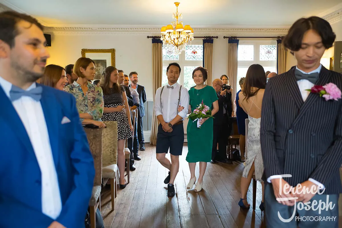 10_County_Arms_Wandsworth_Wedding_Ceremony_Photos Guests seated and standing at a wedding ceremony in a bright room at County Arms Wandsworth. A couple walks down the aisle, with the woman holding a bouquet. A man in a suit is at the front. The room has chandeliers, a large mirror, and several windows with drapes.