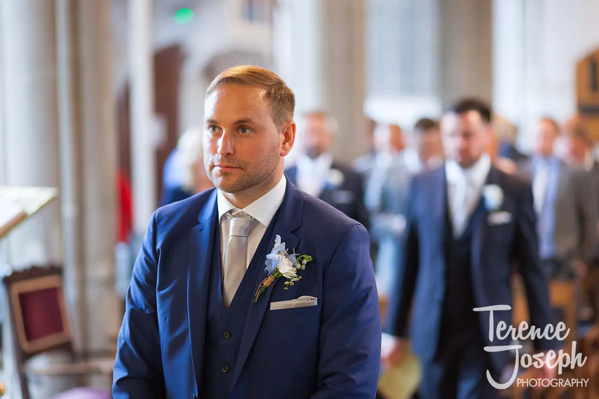 10_St_Andrews_Church_Wedding_Ceremony At a Moreteyne Manor wedding, a groom in a blue suit stands at the altar in a church. With a white boutonniere and tie, he gazes ahead, while the blurred background reveals groomsmen in similar attire.