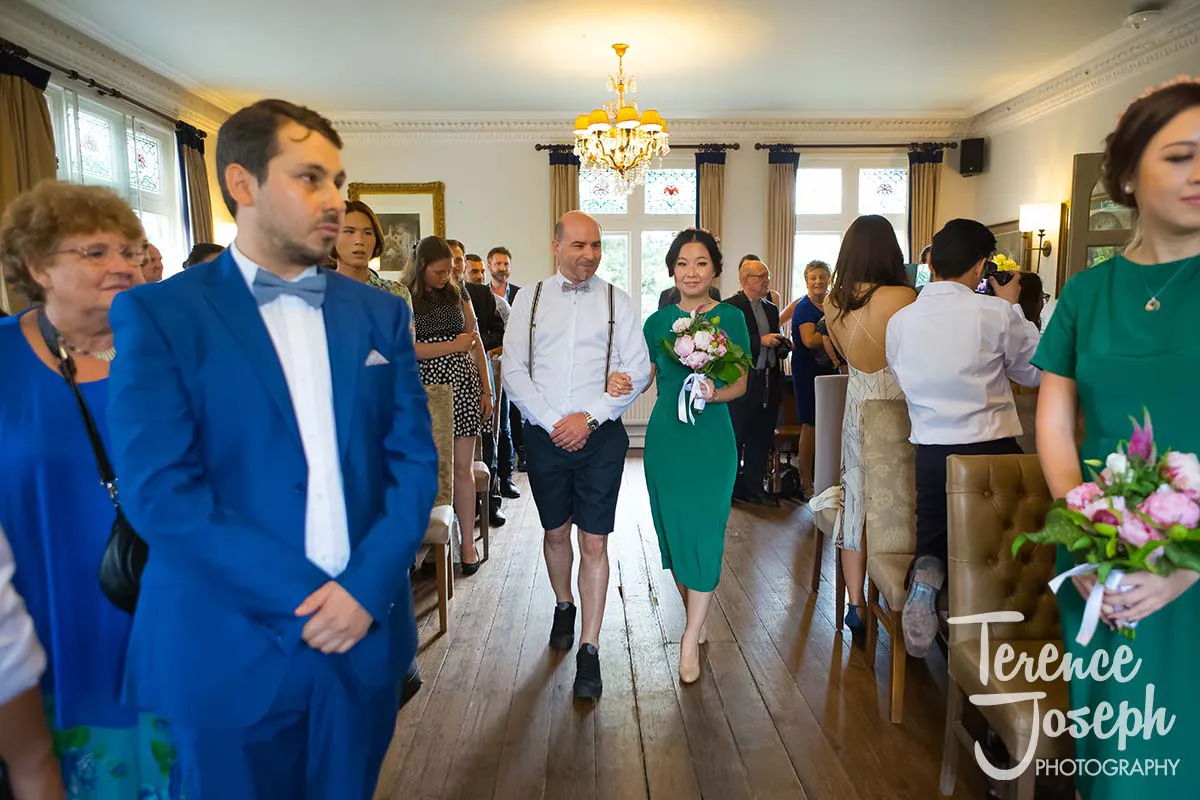 11_County_Arms_Wandsworth_Wedding_Ceremony_Photos A County Arms Wandsworth wedding unfolds in a bright room with guests. A man in shorts and a white shirt escorts a woman in a green dress holding a bouquet down the aisle, under elegant chandeliers and large windows, as attendees watch, some standing, some seated.