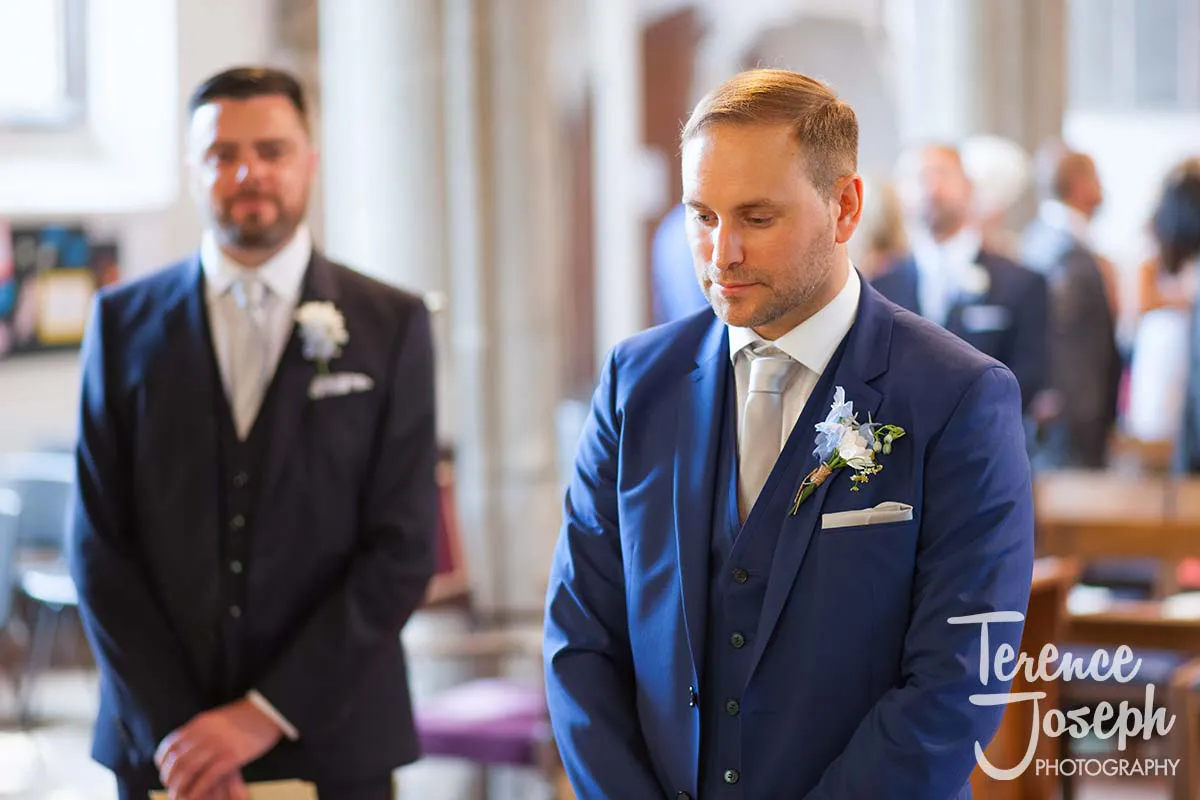 11_St_Andrews_Church_Wedding_Ceremony A groom in a blue suit and silver tie stands indoors at a wedding ceremony, looking down. A groomsman in a navy suit and light tie is visible in the background. Guests are seen slightly blurred in the church setting, captured exquisitely by the Moreteyne Manor Wedding Photographer.