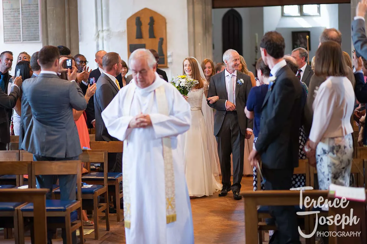 12_St_Andrews_Church_Wedding_Ceremony A bride in a white dress walks down the aisle at Moreteyne Manor, accompanied by an older man. The church echoes with the shutter clicks of guests capturing memories as a priest in white robes leads the ceremony.