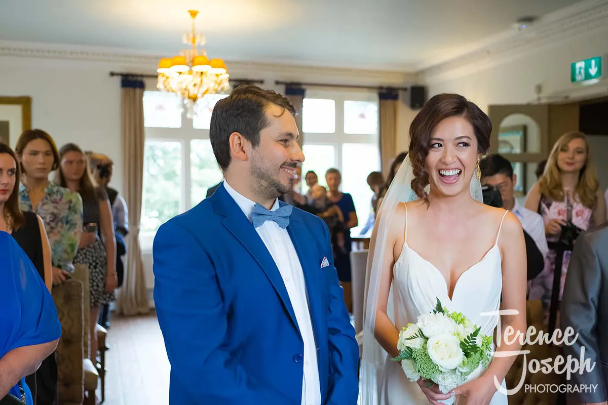 13_County_Arms_Wandsworth_Wedding_Ceremony_Photos A bride and groom stand together at their County Arms Wandsworth wedding ceremony, smiling joyfully. The bride holds a bouquet of white flowers, and the groom wears a blue suit with a bow tie. Guests are seated in the background, and a chandelier hangs overhead.