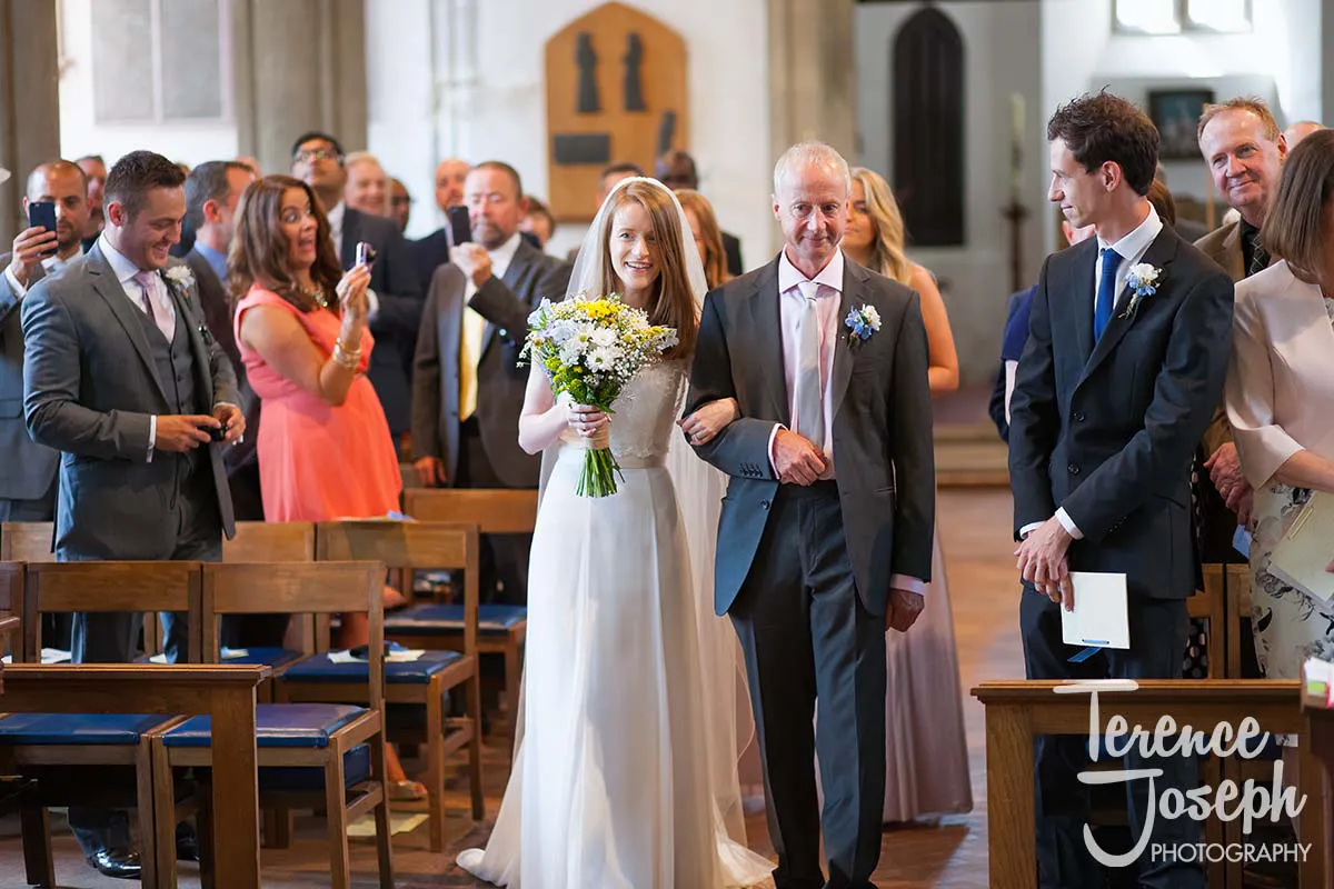 13_St_Andrews_Church_Wedding_Ceremony A bride in a white dress holds a bouquet and walks down the aisle with an older man in a suit, surrounded by smiling guests. At the front, her groom waits eagerly. Captured by a Moreteyne Manor wedding photographer, the vibrant church setting features elegant wooden pews.