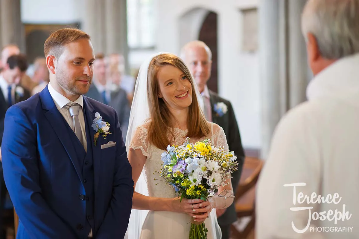 14_St_Andrews_Church_Wedding_Ceremony A bride and groom stand at the altar during their wedding ceremony, captured beautifully by a Moreteyne Manor Wedding Photographer. The bride, smiling warmly, holds a bouquet of white and yellow flowers, while the groom, in a blue suit, looks on. Several guests are visible in the background.