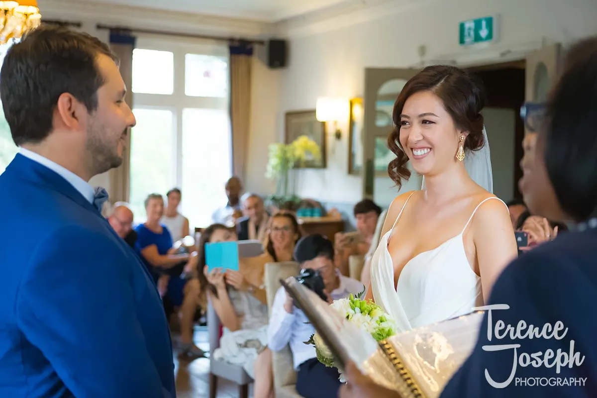15_County_Arms_Wandsworth_Wedding_Ceremony_Photos A bride and groom exchange smiles at their County Arms Wandsworth wedding ceremony. The bride clutches a bouquet as guests capture the moment in photos. Standing beside them, an officiant dressed in black oversees the vows.