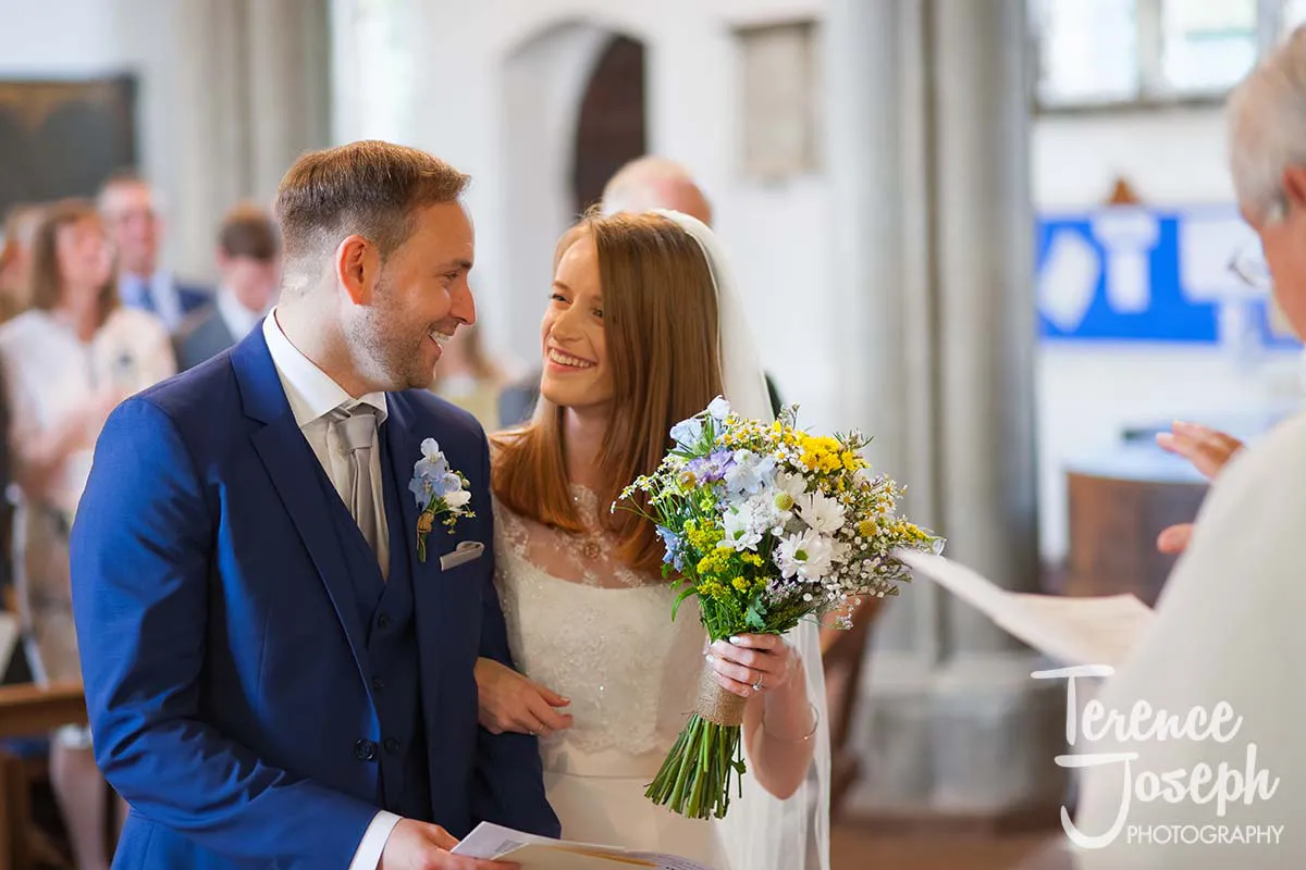 15_St_Andrews_Church_Wedding_Ceremony A smiling couple, dressed formally, stand together inside a church. The woman holds a bouquet of yellow and white flowers. As they face each other happily, surrounded by wedding guests in the background, the moment is perfectly captured by a Moreteyne Manor Wedding Photographer.