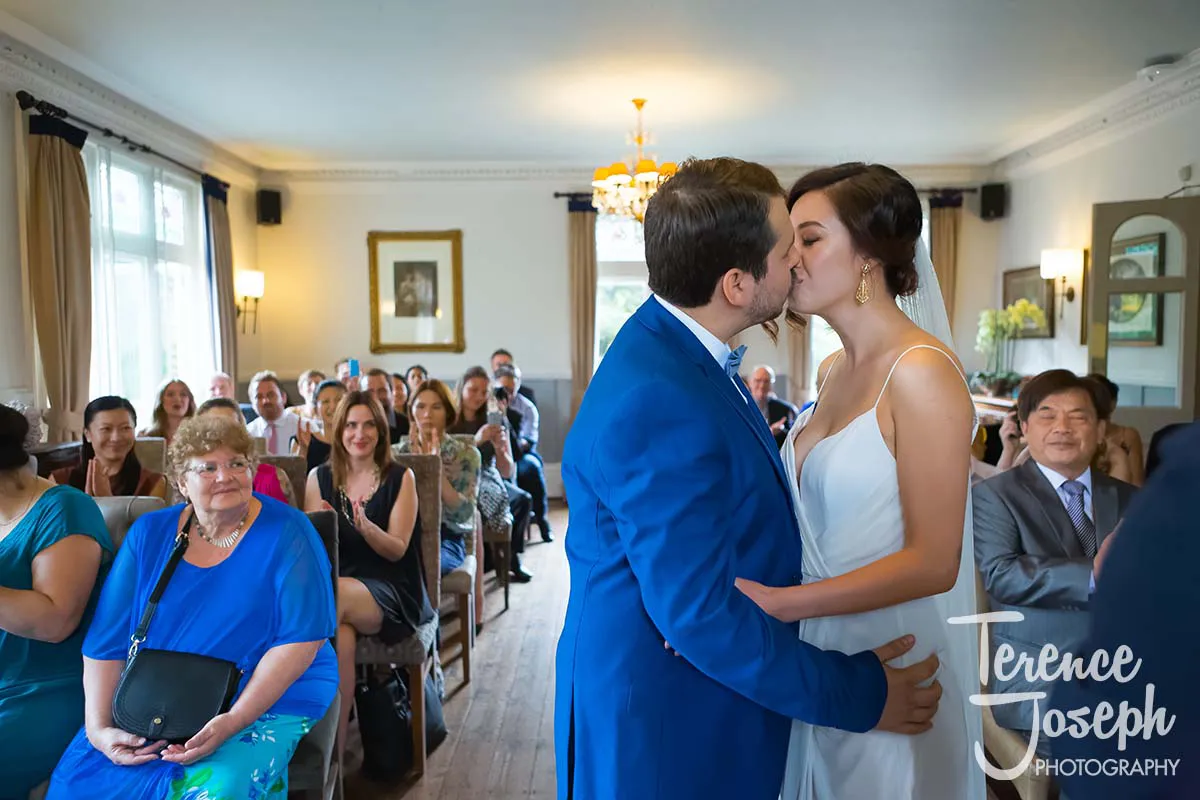 16_County_Arms_Wandsworth_Wedding_Ceremony_Photos At the County Arms Wandsworth, the bride and groom share a tender kiss in a brightly lit room filled with clapping, smiling guests. The bride's white dress and the groom's blue suit complement the elegant chandelier and framed pictures that adorn the walls.