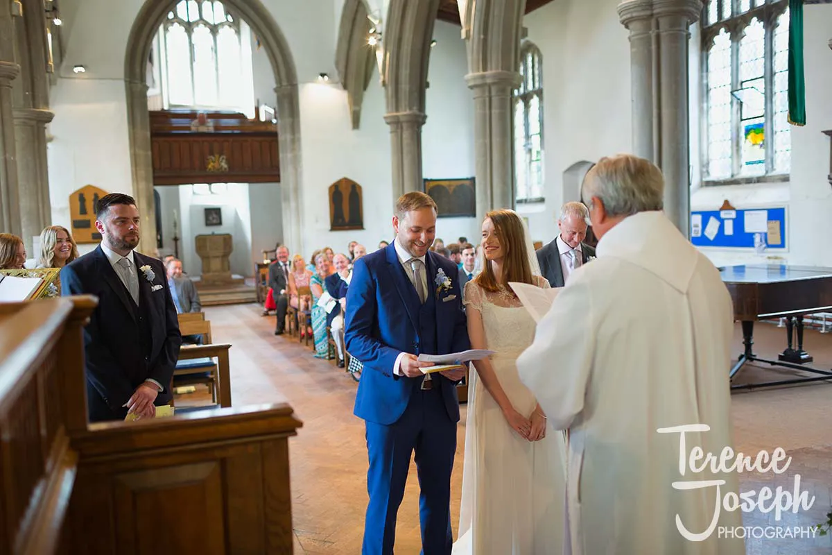16_St_Andrews_Church_Wedding_Ceremony A bride and groom stand at the altar in a church, smiling as they exchange vows. A priest in white robes leads the ceremony, while guests seated on wooden pews watch attentively. The scene is beautifully captured by a Moreteyne Manor Wedding Photographer amidst tall arches and stained glass windows.