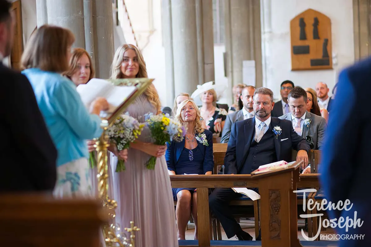 17_St_Andrews_Church_Wedding_Ceremony A wedding ceremony scene in a church, captured by a Moreteyne Manor wedding photographer, features attendees seated and standing. A woman reads from a lectern while another holds a bouquet. The groom sits in the front, flanked by family members, amidst a solemn and festive atmosphere.