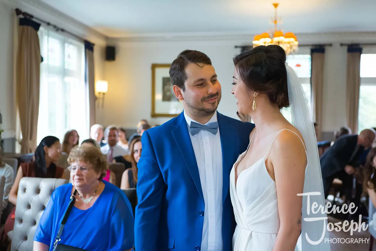 18_County_Arms_Wandsworth_Wedding_Ceremony_Photos At their County Arms Wandsworth wedding, a bride and groom stand facing each other, smiling in a well-lit room with guests seated behind them. The groom wears a blue suit and bow tie, while the bride is in a white dress. Large windows and elegant decor enhance the joyous atmosphere.
