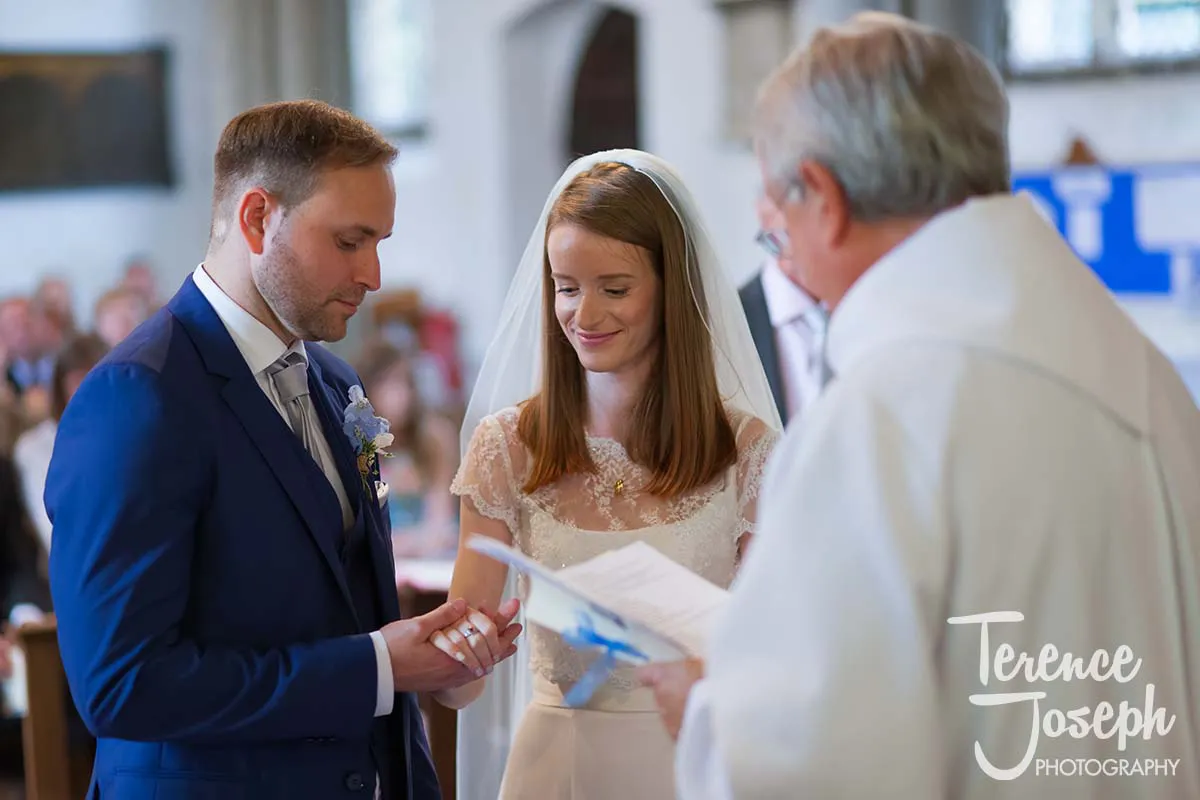18_St_Andrews_Church_Wedding_Ceremony Amidst the elegance of Moreteyne Manor, a bride and groom exchange rings during their church ceremony. The groom dons a blue suit, while the bride dazzles in her white dress and veil. A priest stands before them, paper in hand, perfectly captured by the wedding photographer.