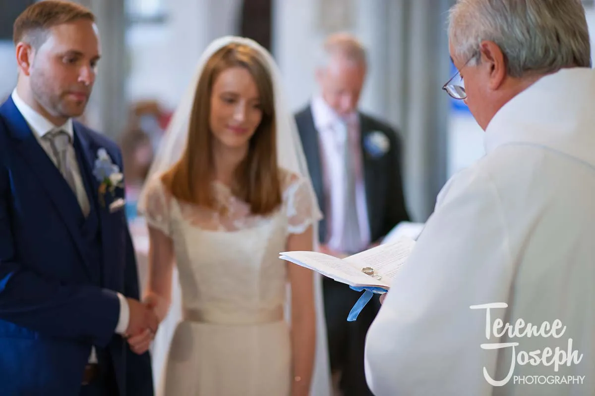 19_St_Andrews_Church_Wedding_Ceremony A bride and groom stand before the priest, who is holding an open book at their Moreteyne Manor wedding ceremony. The groom dons a blue suit, and the bride shines in her white dress, while their special day is beautifully captured by a skilled wedding photographer. A man is blurred in the background.