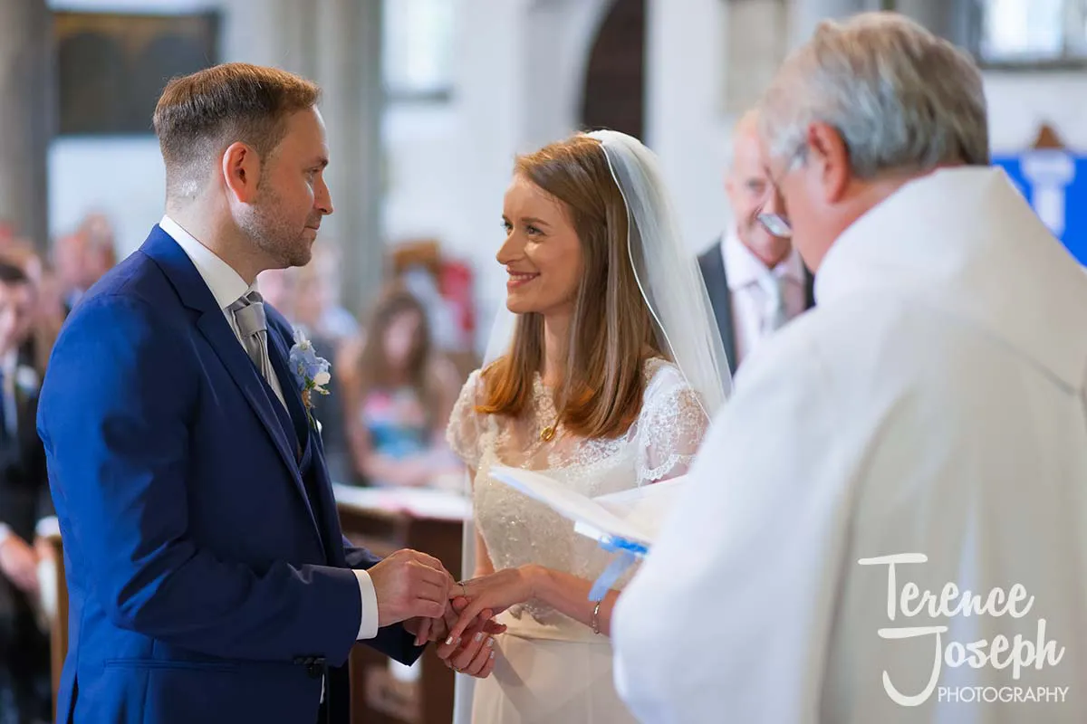 20_St_Andrews_Church_Wedding_Ceremony During the joyful and intimate church ceremony, a Moreteyne Manor wedding photographer captures the magical moment as the bride in her white dress and veil exchanges rings with the groom in his blue suit, while a priest reads from a book nearby.