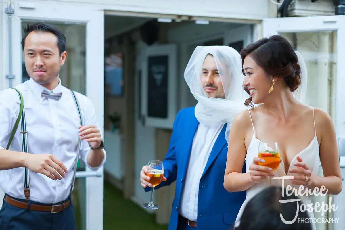 21_County_Arms_Wandsworth_Wedding_Breakfast_Photos At a charming County Arms Wandsworth wedding, a smiling bride in a white dress holds a drink. Beside her, a man in a blue suit with a veil over his head shares the joy with his own drink. Nearby, another man in a white shirt and bow tie gestures animatedly.