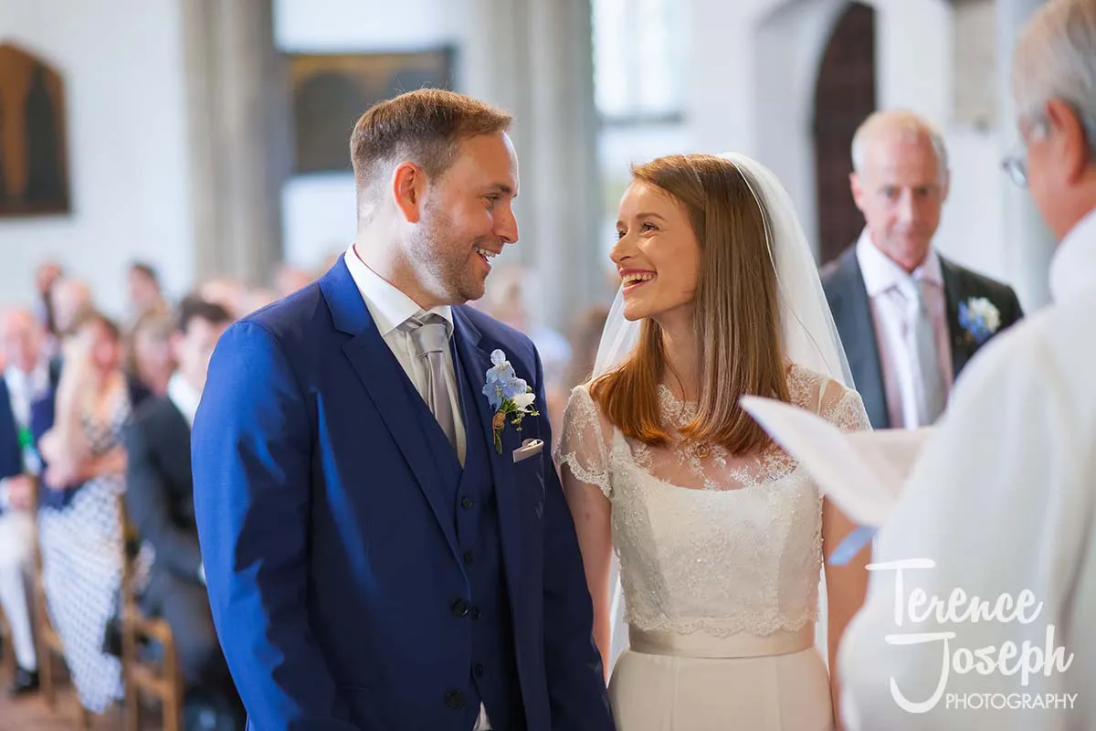 21_St_Andrews_Church_Wedding_Ceremony A bride and groom smile at each other during their Moreteyne Manor wedding ceremony. The groom is wearing a blue suit with a white shirt, and the bride is in a white lace dress. Guests are seated in the background, and a priest is holding a paper.
