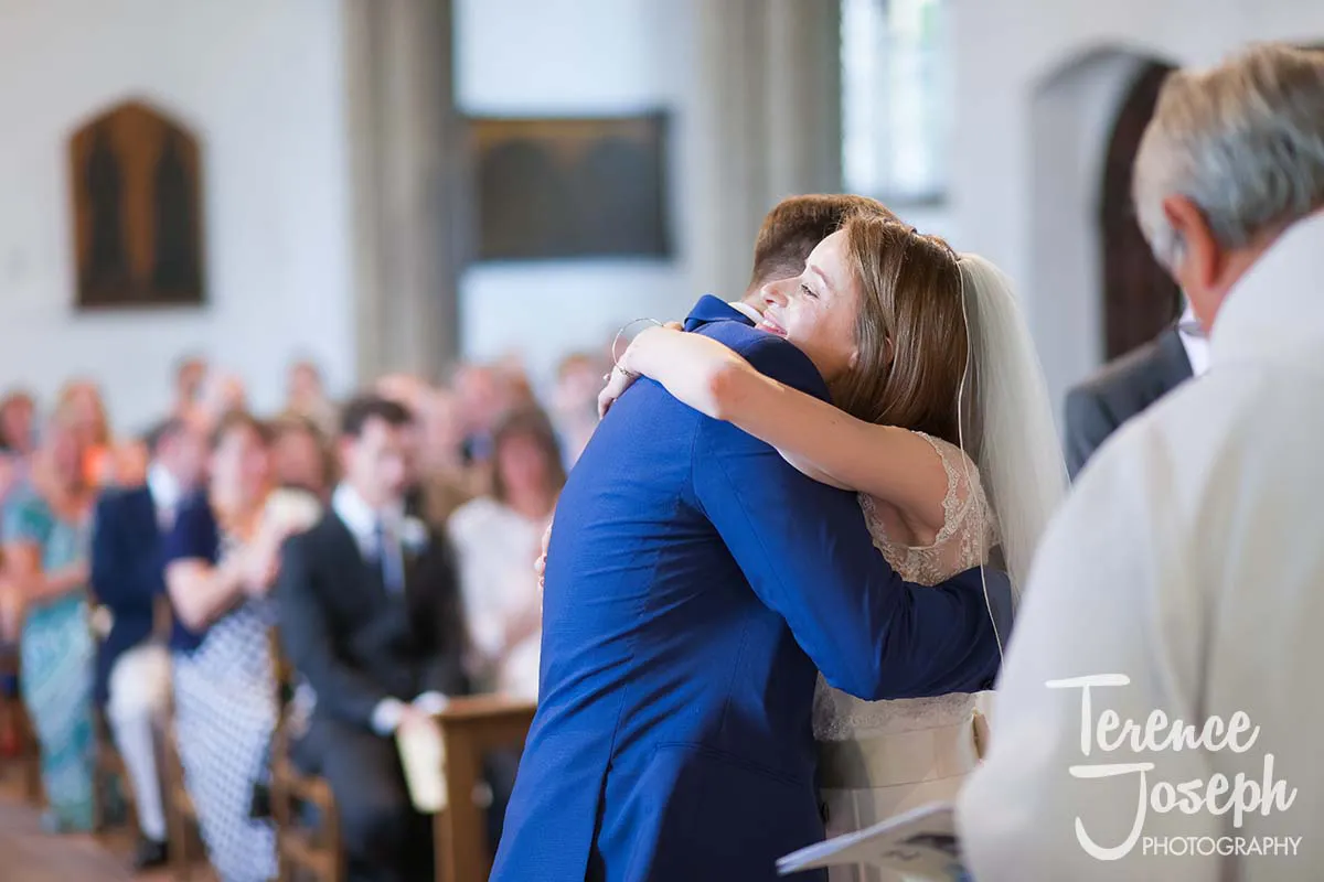22_St_Andrews_Church_Wedding_Ceremony A bride and groom warmly embrace during their wedding ceremony in a church, beautifully captured by a Moreteyne Manor wedding photographer. The bride wears a white dress and veil, while the groom dons a blue suit. Guests sit in the background, witnessing their special moment.