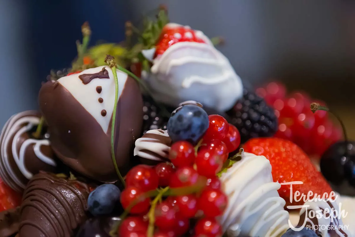 23_County_Arms_Wandsworth_Wedding_Breakfast_Photos A close-up of assorted chocolate-dipped strawberries, including one decorated to resemble a tuxedo, surrounded by blueberries, redcurrants, and blackberries—perfect for a County Arms Wandsworth wedding. The photo features the logo "Terence Joseph Photography" in the corner.