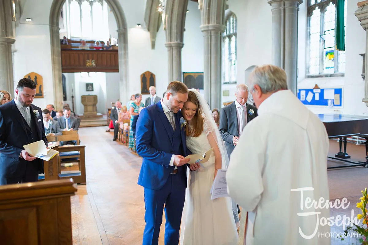 23_St_Andrews_Church_Wedding_Ceremony A bride and groom stand smiling, holding a program inside a bright church. Nearby, a priest in white robes offers blessings. Guests seated in pews witness the joy, as a man in a blue suit stands prominently, capturing moments like a Moreteyne Manor wedding photographer would.