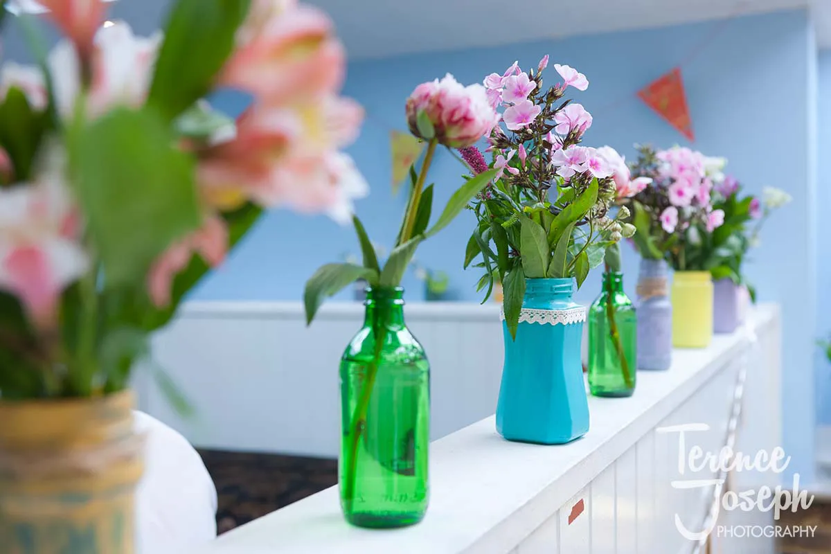 24_County_Arms_Wandsworth_Wedding_Breakfast_Photos Colorful flowers arranged in glass bottles, including green and blue, adorn a white ledge at a County Arms Wandsworth wedding. The light blue backdrop features decorative triangle banners hanging above.