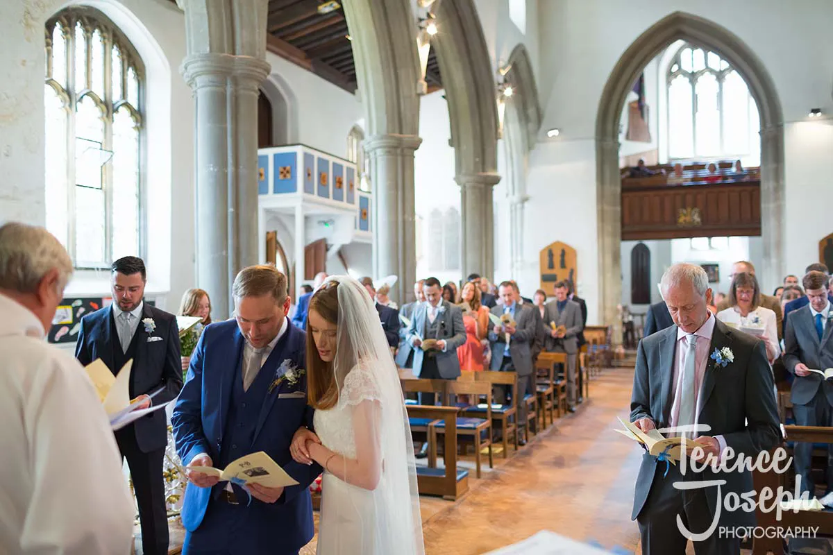 24_St_Andrews_Church_Wedding_Ceremony A wedding ceremony unfolds in a church with the bride in a white gown and veil beside the groom, both clutching programs. Guests in formal attire observe the proceedings. Tall arches and stained glass windows adorn the scene, reminiscent of a Moreteyne Manor Wedding Photographer's exquisite touch.