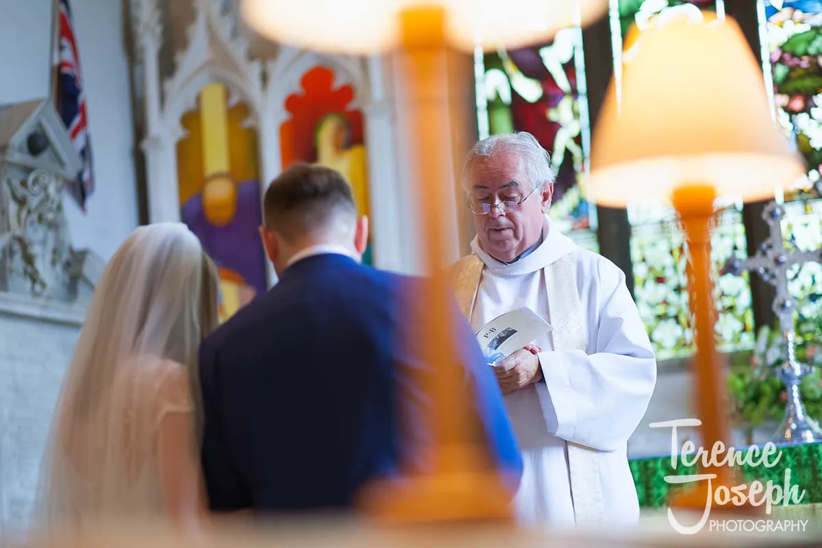 25_St_Andrews_Church_Wedding_Ceremony Inside the church, a priest officiates a wedding ceremony captured elegantly by a Moreteyne Manor Wedding Photographer. The bride, wearing a veil, and groom stand facing him, as stained glass windows and classic decor create a stunning backdrop.