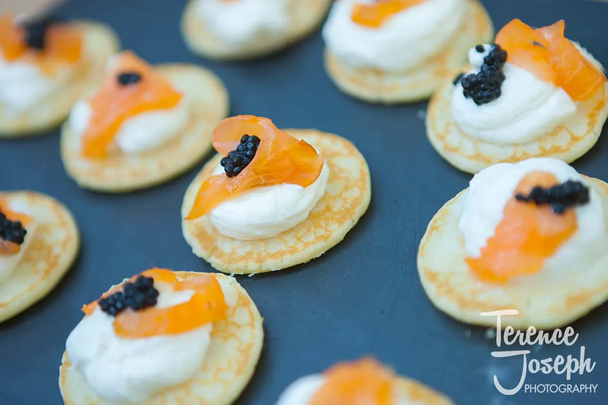 26_County_Arms_Wandsworth_Wedding_Breakfast_Photos Gourmet appetizers at a County Arms Wandsworth wedding feature small pancakes topped with creamy spread, slices of smoked salmon, and a dollop of black caviar, elegantly arranged on a dark serving platter.