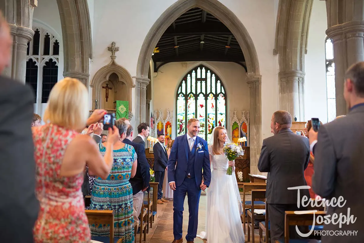 26_St_Andrews_Church_Wedding_Ceremony A bride and groom, smiling and holding hands, walk down the aisle of a church. Guests are standing and taking photos, capturing every moment like a seasoned Moreteyne Manor Wedding Photographer would. The church is adorned with stained glass windows and wooden beams.