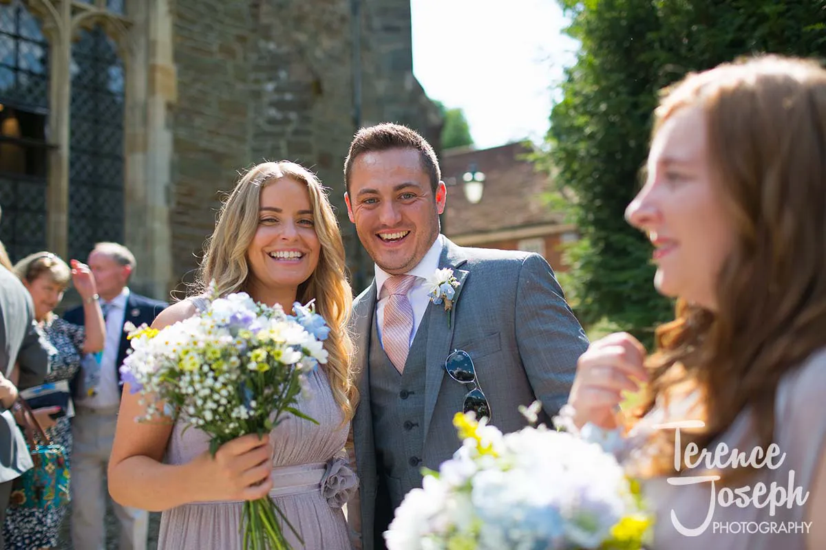27_St_Andrews_Church_Wedding_Ceremony A smiling couple, elegantly dressed and holding flower bouquets, pose outdoors at a Moreteyne Manor wedding. Other guests mingle in the background as the historic building's charm shines under a sunny sky, perfectly captured by their talented photographer.