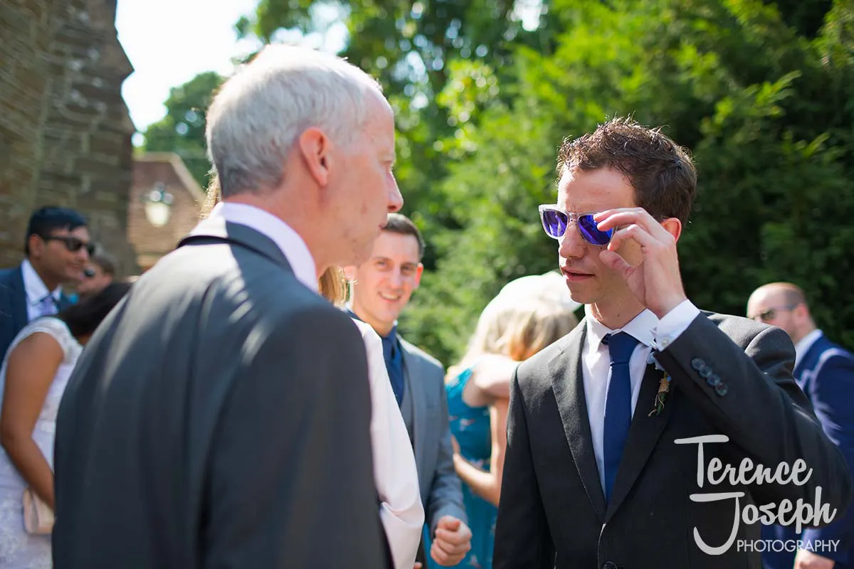 28_St_Andrews_Church_Wedding_Ceremony A man in a suit adjusts his purple sunglasses while engaging in conversation with an older gentleman in a gray suit at an outdoor event. Capturing the essence of Moreteyne Manor wedding moments, guests in formal attire enjoy the sunlit backdrop.