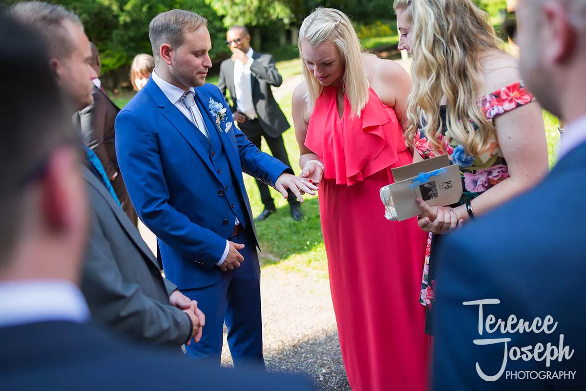 29_St_Andrews_Church_Wedding_Ceremony A groom in a blue suit proudly displays his wedding ring to an admiring group under the sunny skies. The Moreteyne Manor Wedding Photographer captures a woman in a red dress appreciating the ring amidst elegantly dressed onlookers in the lush, green surroundings.