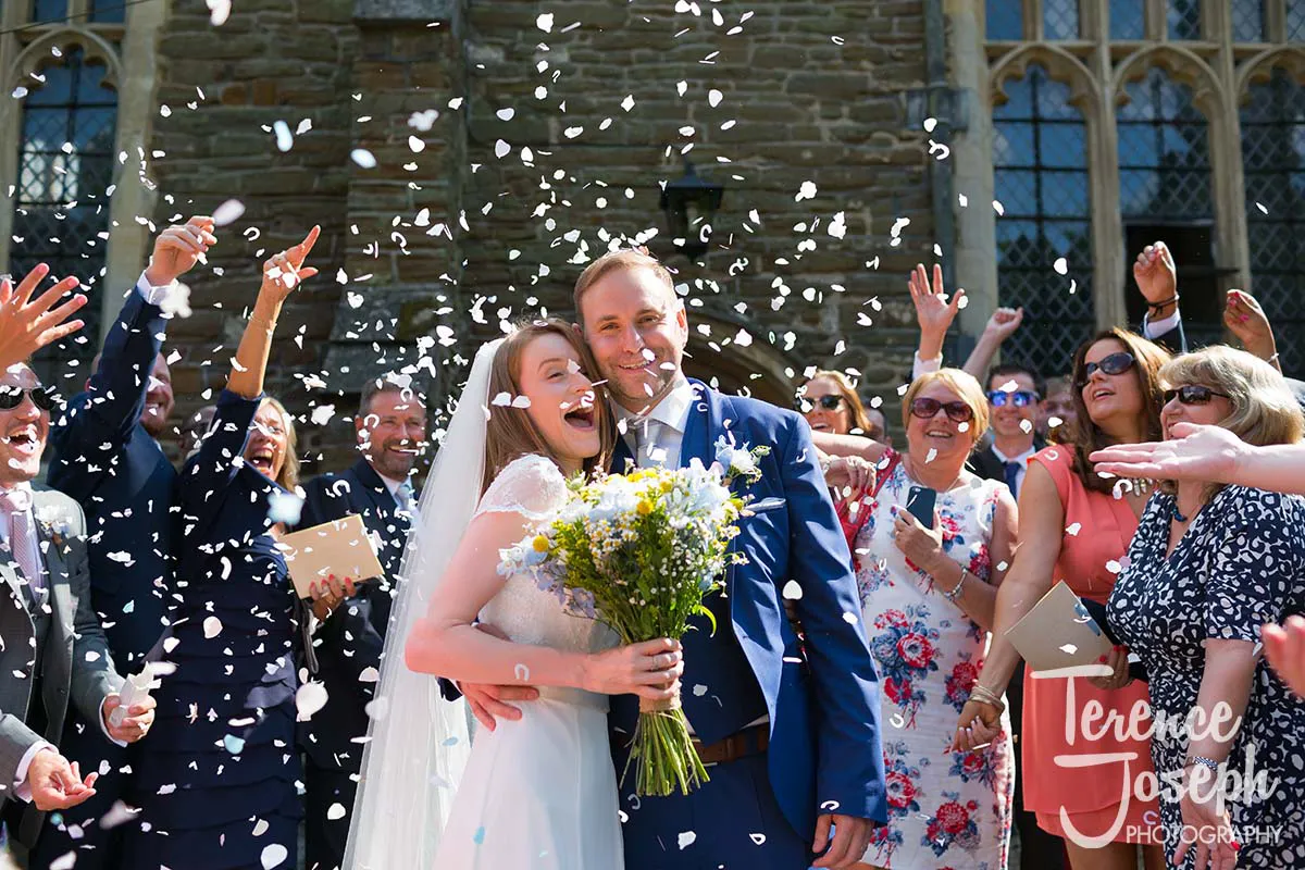 30_St_Andrews_Church_Wedding_Ceremony A joyful Moreteyne Manor wedding scene captures the bride and groom smiling and embracing as confetti swirls around them. The delighted bride clutches a bouquet of flowers while friends and family cheer in celebration, framed by the grandeur of the historic stone building.