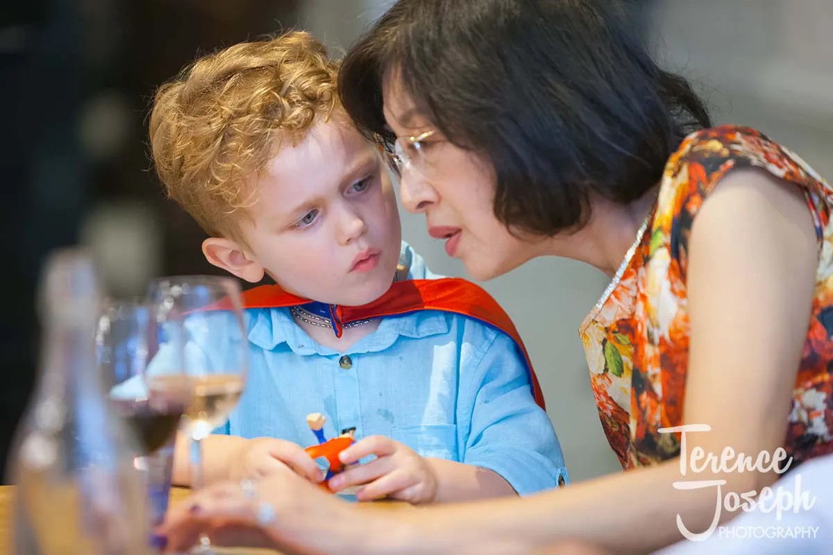 31_County_Arms_Wandsworth_Wedding_Breakfast_Photos A young child in a blue shirt and red cape holds a toy and looks at a woman with glasses. Sitting by a table with wine at the County Arms Wandsworth wedding, the woman leans in to chat with him, creating a moment filled with warmth and joy amid the celebration.