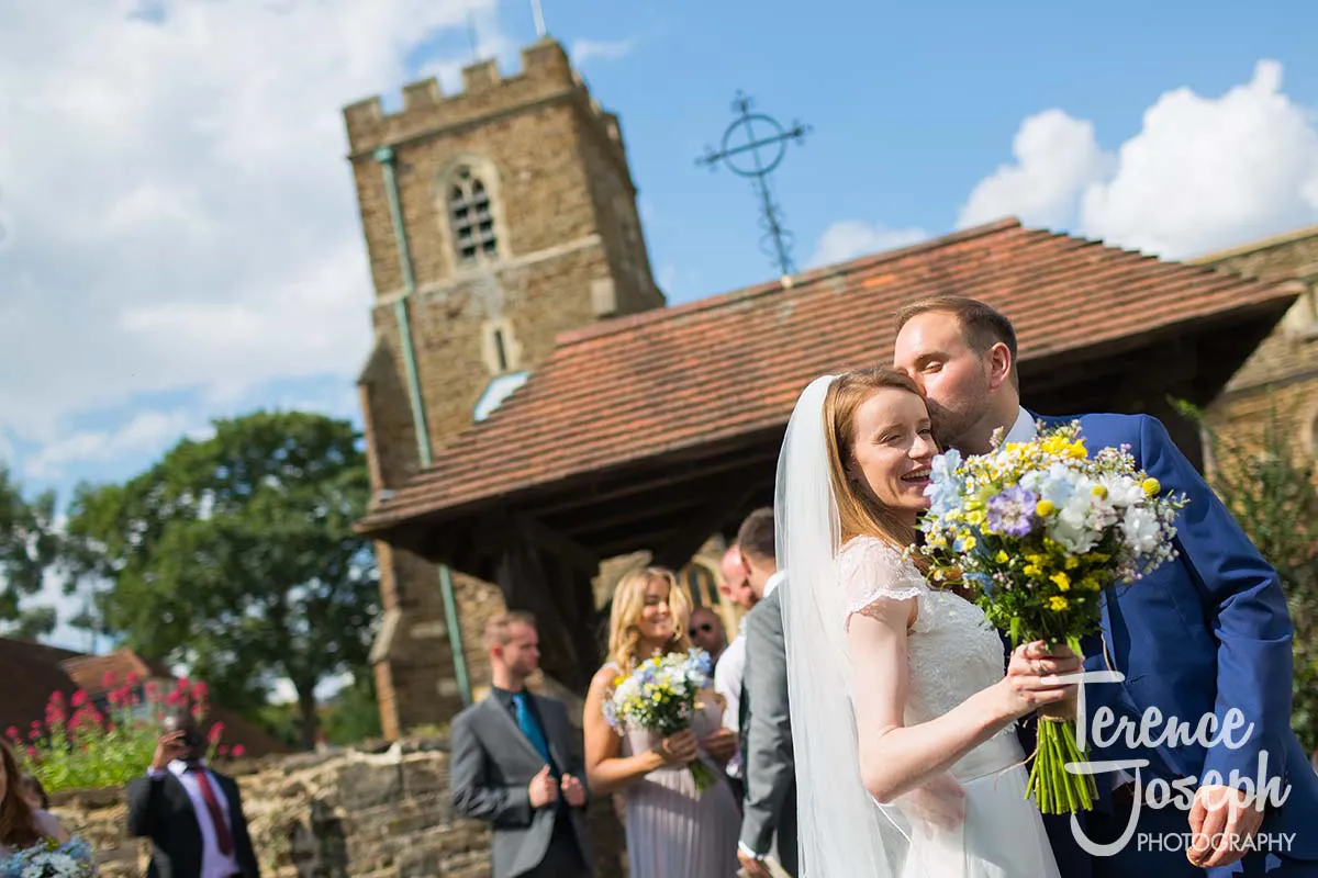 31_St_Andrews_Church_Wedding_Ceremony A bride and groom pose joyfully outside a church on a sunny day, captured perfectly by Moreteyne Manor Wedding Photographer. The bride holds a bouquet of yellow and white flowers as the groom kisses her forehead, while guests chat and smile in the background.
