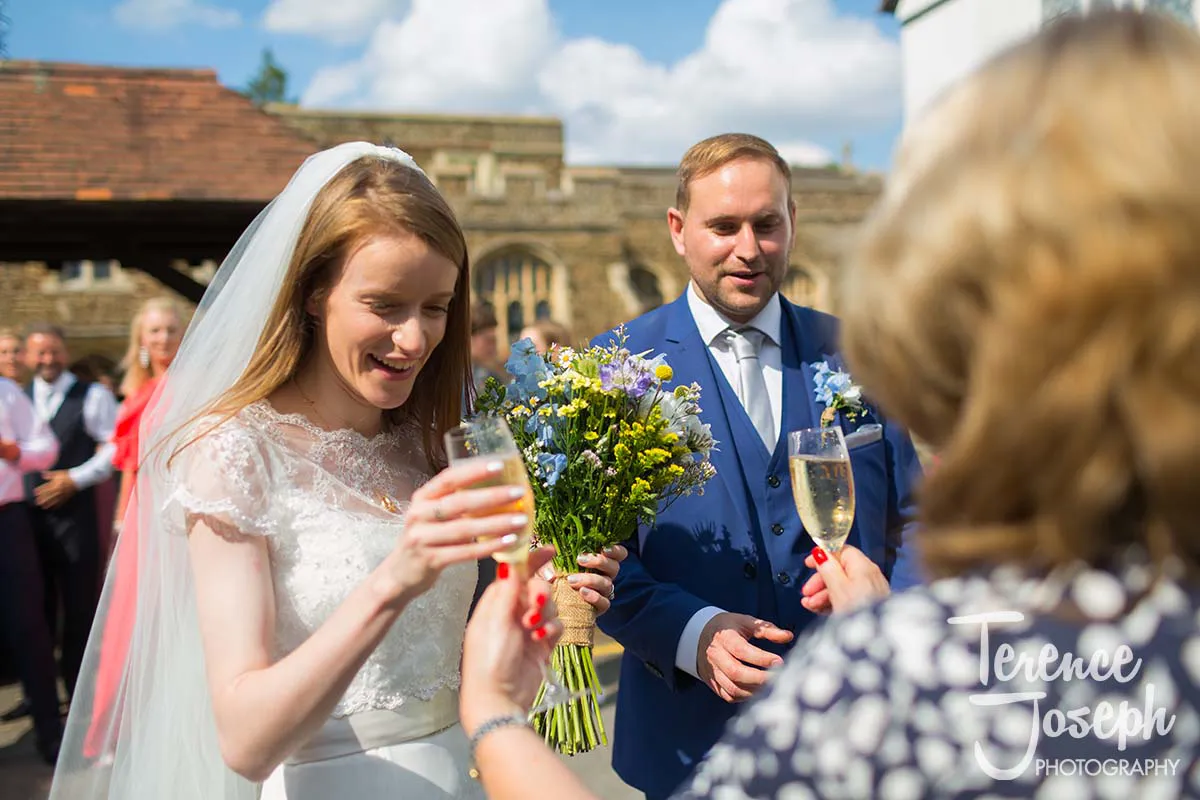32_St_Andrews_Church_Wedding_Ceremony A bride in a white dress and veil holds a drink, smiling, next to a groom in a blue suit with a flower bouquet. Captured by the talented Moreteyne Manor Wedding Photographer, they stand outside as guests mingle under the sunny sky. A woman offers the bride another glass.