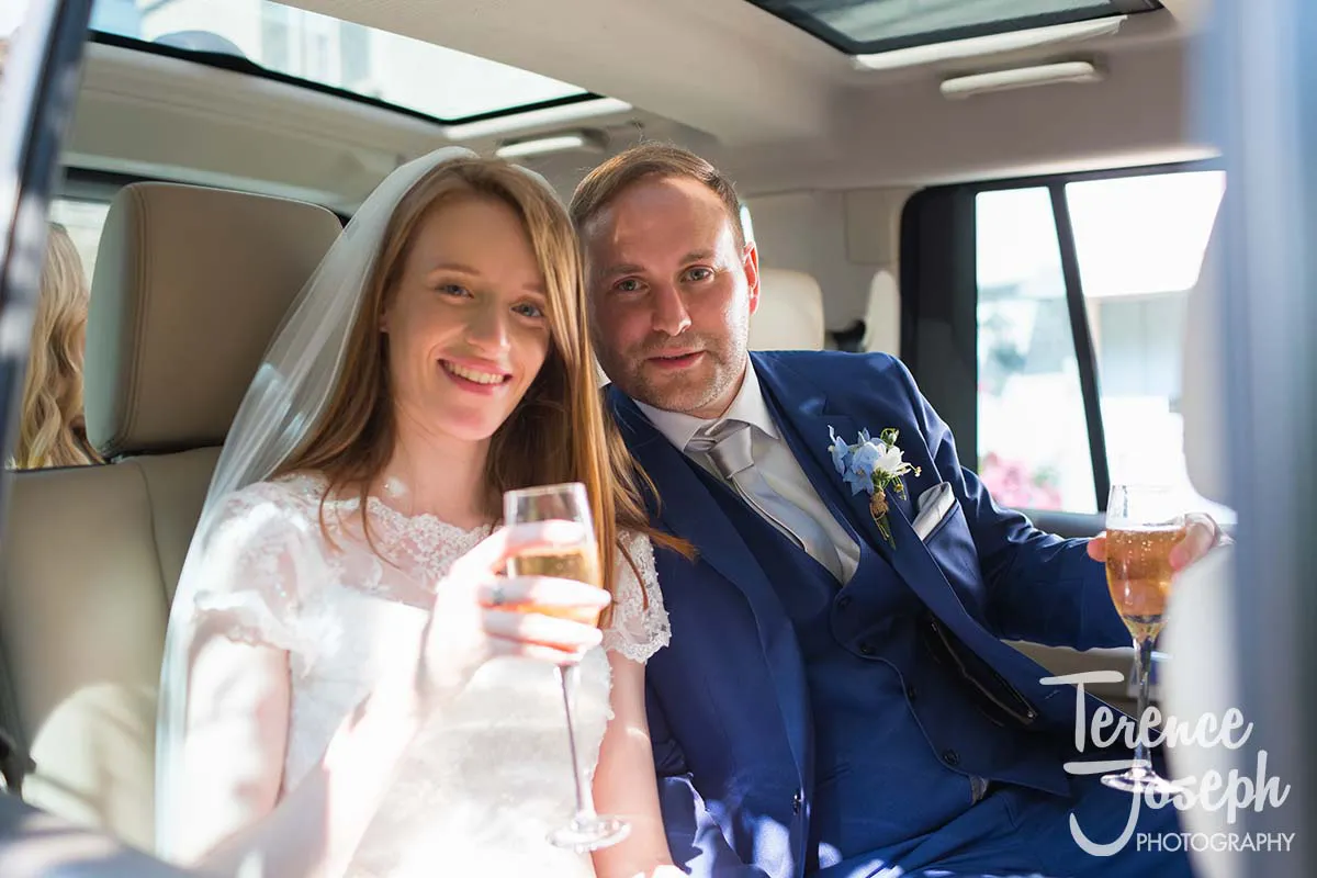 33_St_Andrews_Church_Wedding_Ceremony A bride and groom, captured by a Moreteyne Manor wedding photographer, smile joyfully while sitting in a car. The bride, in a white dress and veil, holds a wine glass. The groom, donned in a blue suit with a boutonniere, also holds a drink. They exude relaxation and happiness.