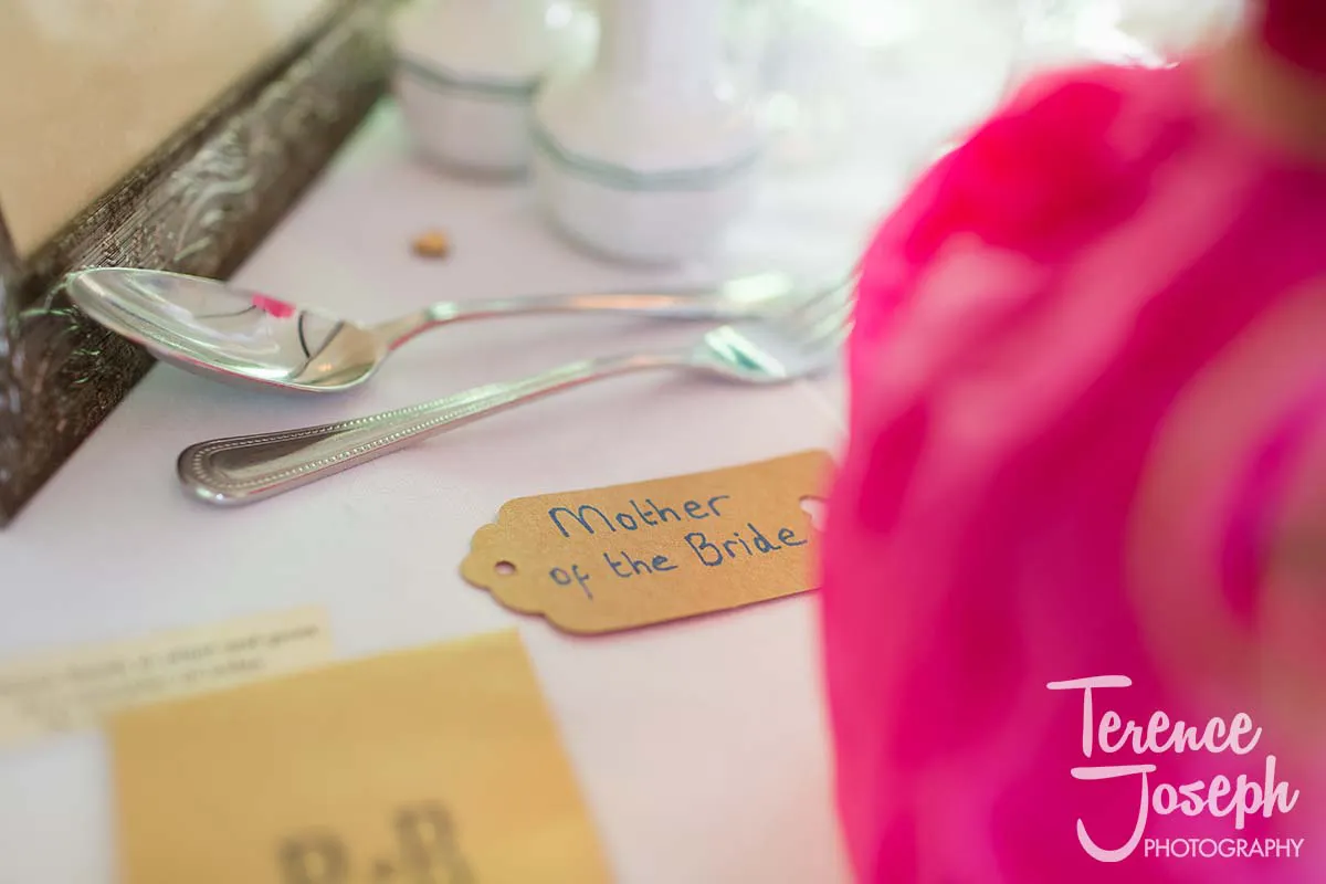 34_Moreteyne_Manor_Wedding_Breakfast A close-up of a wedding place setting at Moreteyne Manor shows a handwritten tag reading "Mother of the Bride." Silverware gleams beside it, with a blurred pink object in the foreground. The image is elegantly signed "Terence Joseph Photography" in the corner.